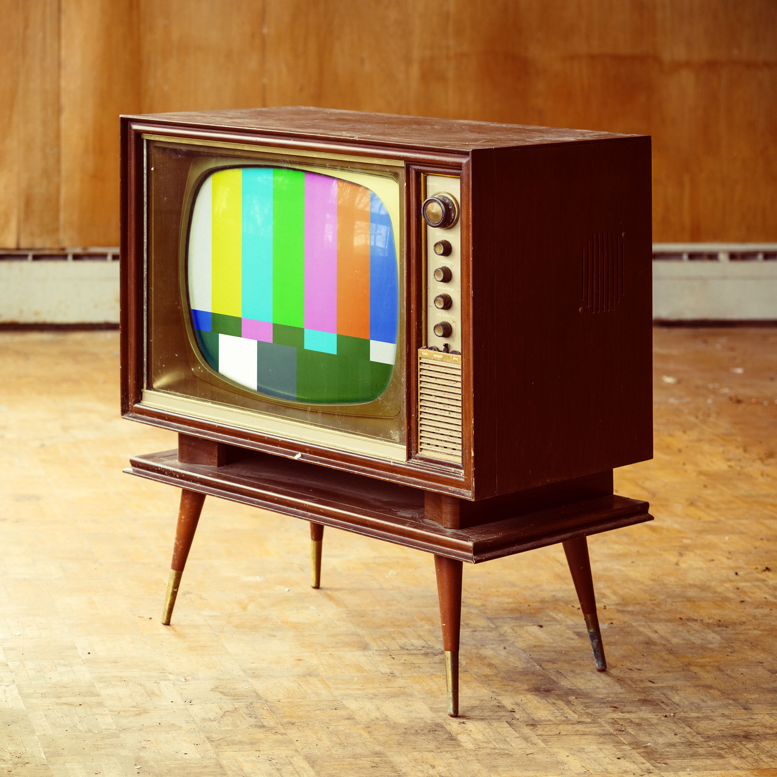 Vintage wooden console TV turned on with color block screen on a wooden floor with a wooden wall in the background