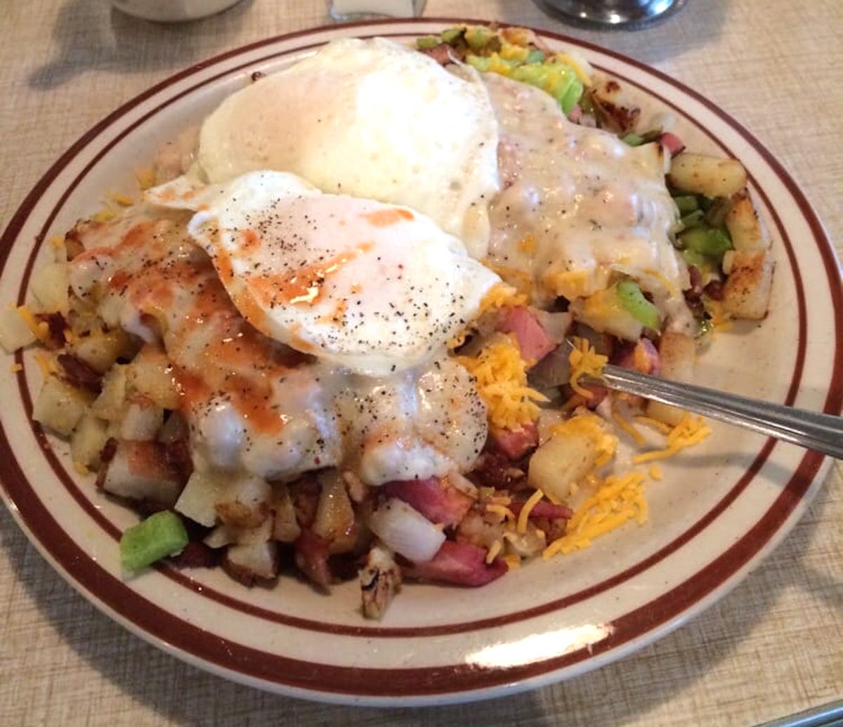 A plate of breakfast hash with diced potatoes, ham, melted cheese, chopped green peppers, and two eggs on top, covered in gravy and topped with hot sauce, with a fork on the side.
