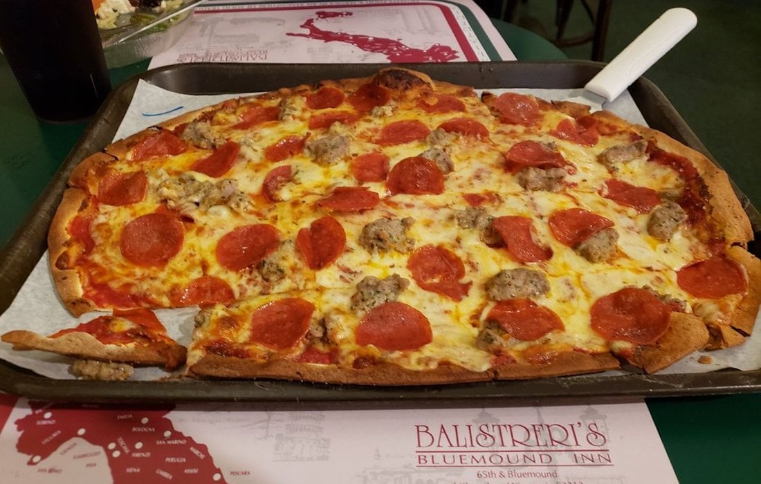 A rectangular pizza topped with pepperoni and sausage sits on a metal tray with a serving spatula. One corner slice is partially removed. The pizza is on a table with a placemat labeled "Balistreri's Bluemound Inn.