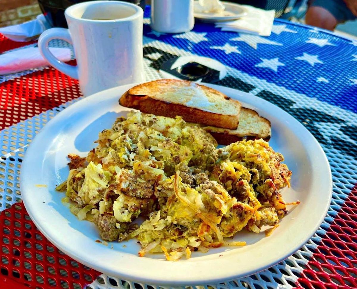 A plate with scrambled eggs, ground sausage, and hash browns, served with two slices of toasted bread and a mug of coffee on a table covered with an American flag tablecloth.