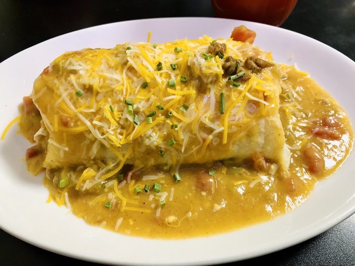 A burrito topped with melted shredded cheese, chopped chives, and smothered in a creamy yellow sauce sits on a white plate against a dark background.