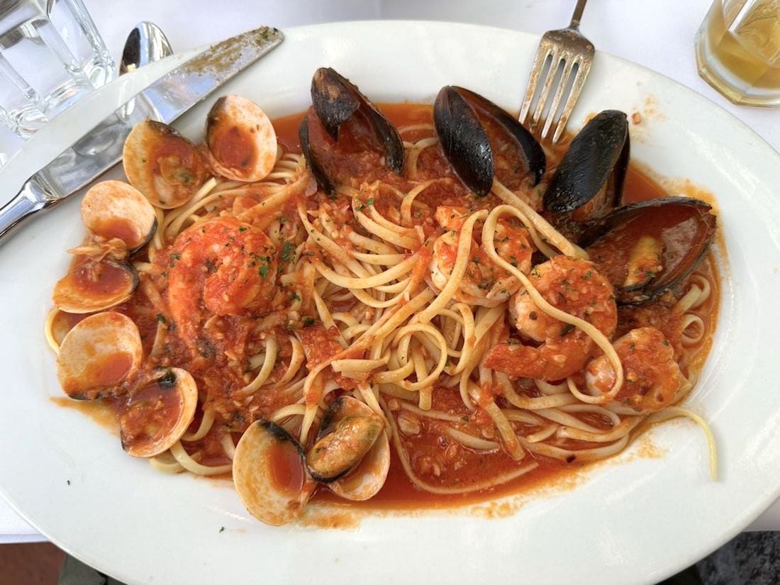 A white plate of linguine pasta with tomato sauce, mussels, clams, and shrimp. A fork and knife rest on the plate’s edge, and a glass of water and a drink are partially visible in the background.