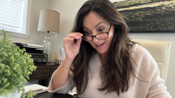A woman with long brown hair and glasses looks surprised while reading at her desk, surrounded by office items like a lamp, printer, papers, and a plant.