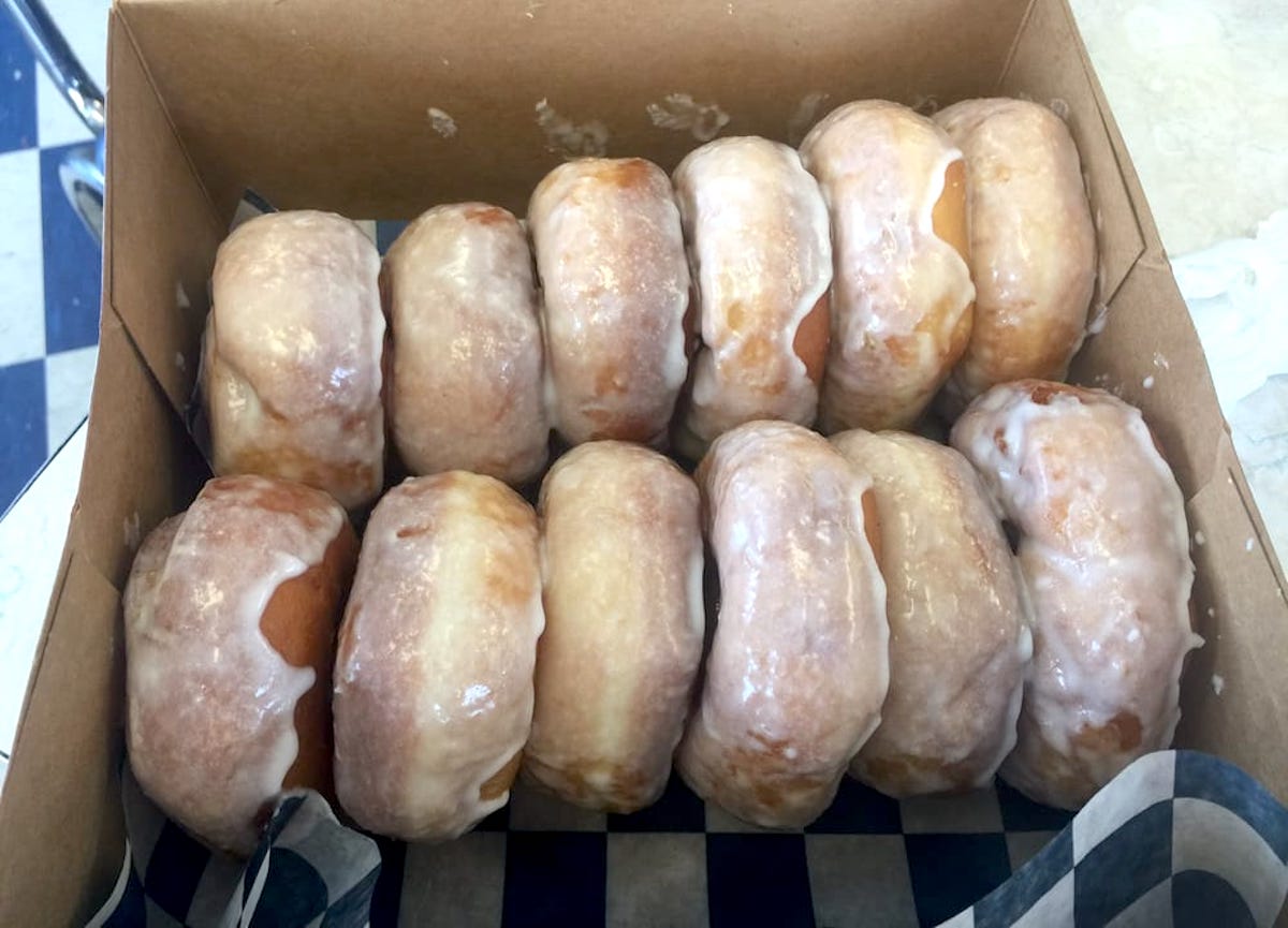 A box of ten glazed donuts arranged in two rows, each donut covered in a shiny sugar glaze. The donuts are in a cardboard box with a blue and white checkered paper lining.