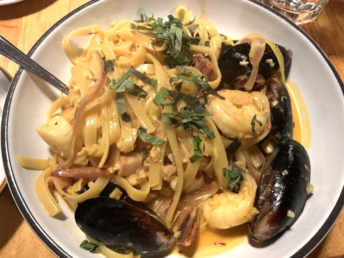 A bowl of seafood pasta with shrimp, mussels, and fettuccine noodles in a creamy sauce, garnished with fresh basil, with a fork on the side and a glass of water partially visible in the background.