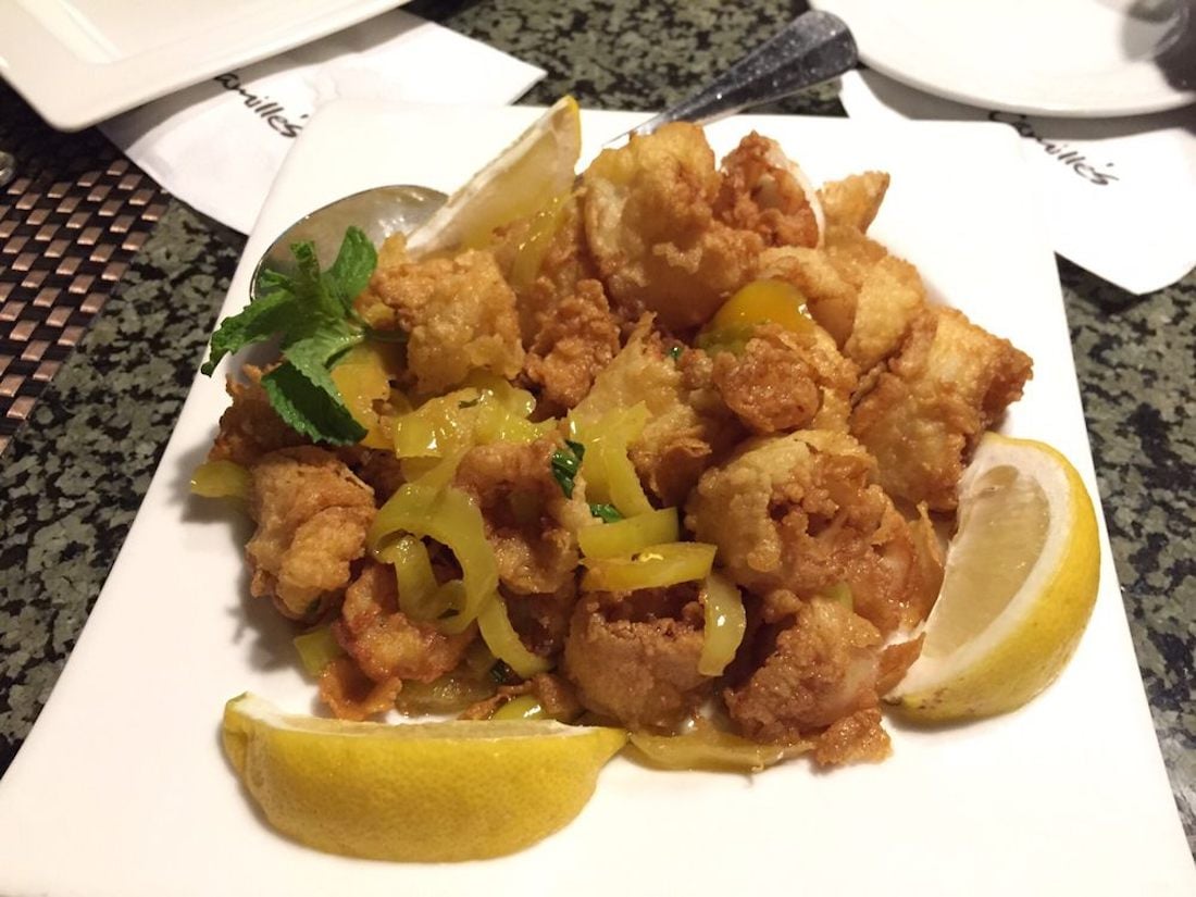 A white plate of fried calamari garnished with yellow peppers, parsley, and lemon wedges, with a serving spoon and napkin in the background.