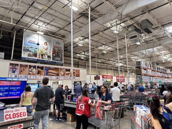 Shoppers wait in line at a busy warehouse store checkout area. Several checkout lanes are open, with carts full of groceries, and a &ldquo;Sorry, this lane CLOSED&rdquo; sign is visible in the foreground.