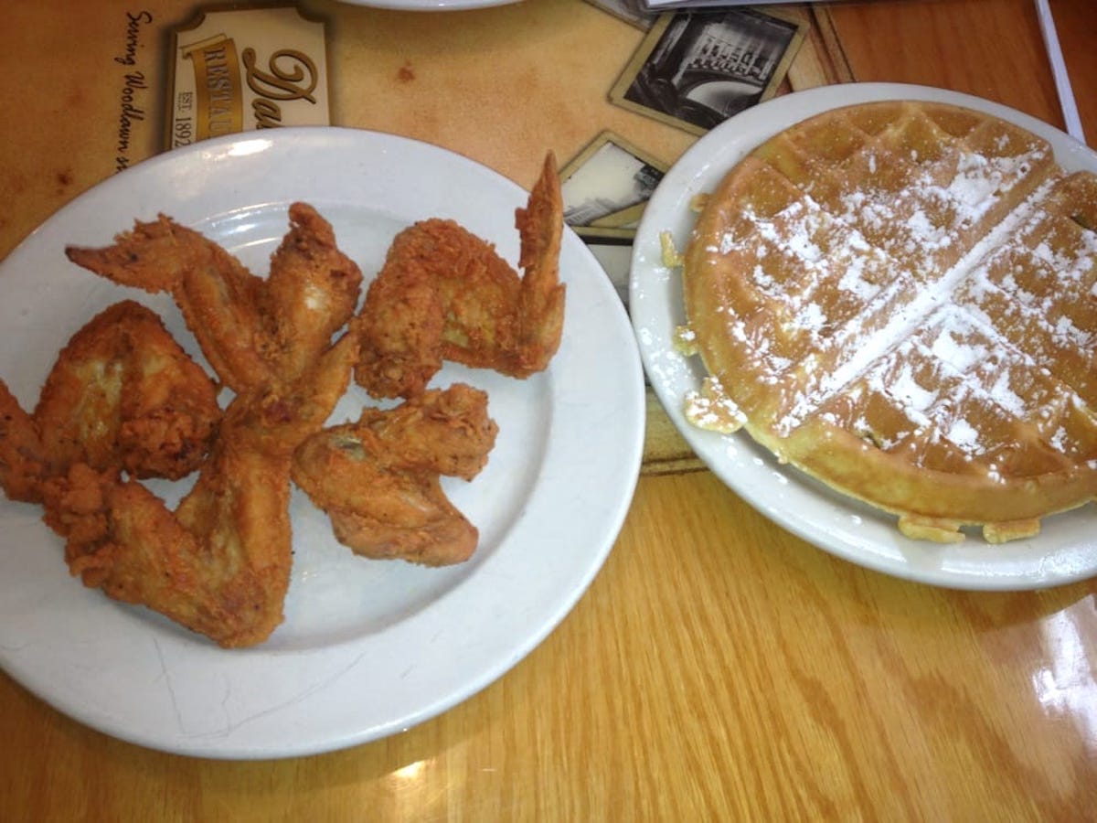 A plate of four fried chicken wings sits next to a plate with a large, round waffle dusted with powdered sugar on a wooden table.