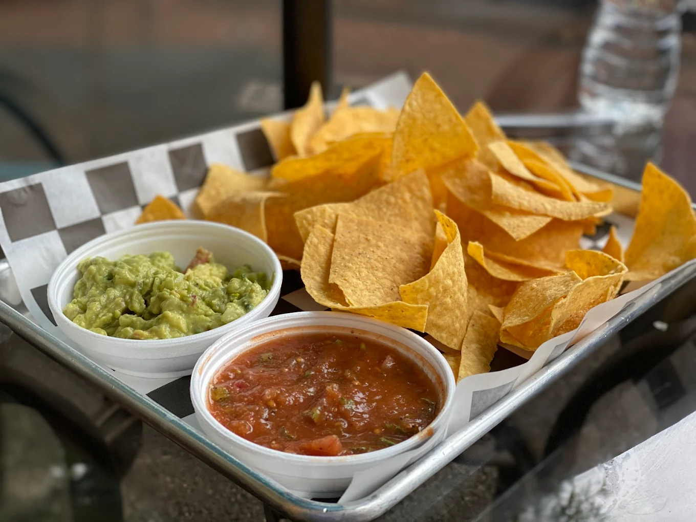 A tray with tortilla chips served alongside small containers of guacamole and salsa, set on a checkered paper liner. A bottle of water is visible in the background.
