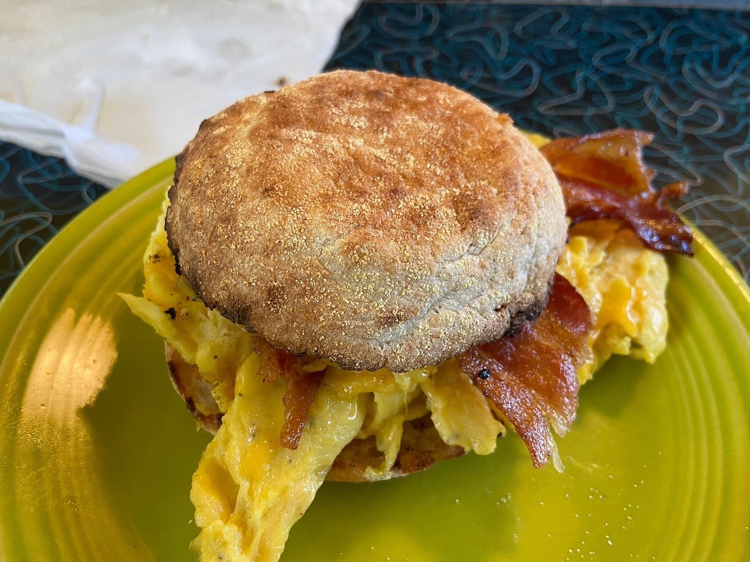 A breakfast sandwich with scrambled eggs and crispy bacon on an English muffin sits on a green plate.