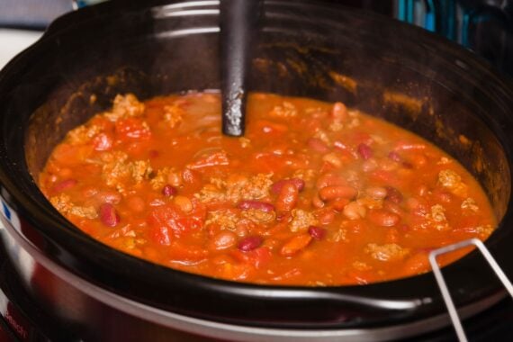 A close-up of chili with beans, tomatoes, and ground meat cooking in a black slow cooker, with a ladle resting inside. Steam rises from the hot, thick mixture.
