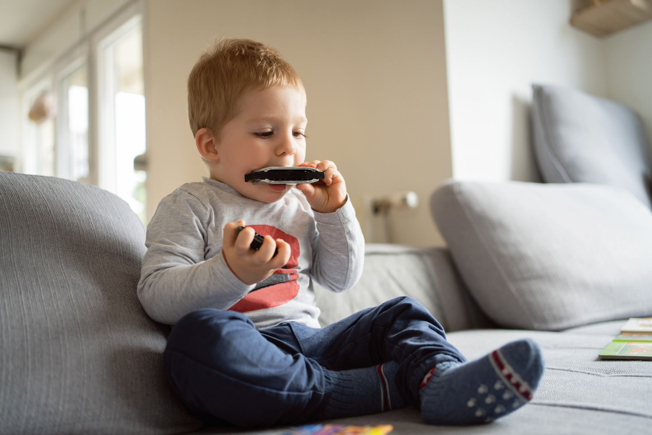 Curious toddler boy sitting on the sofa and blowing the harmonica