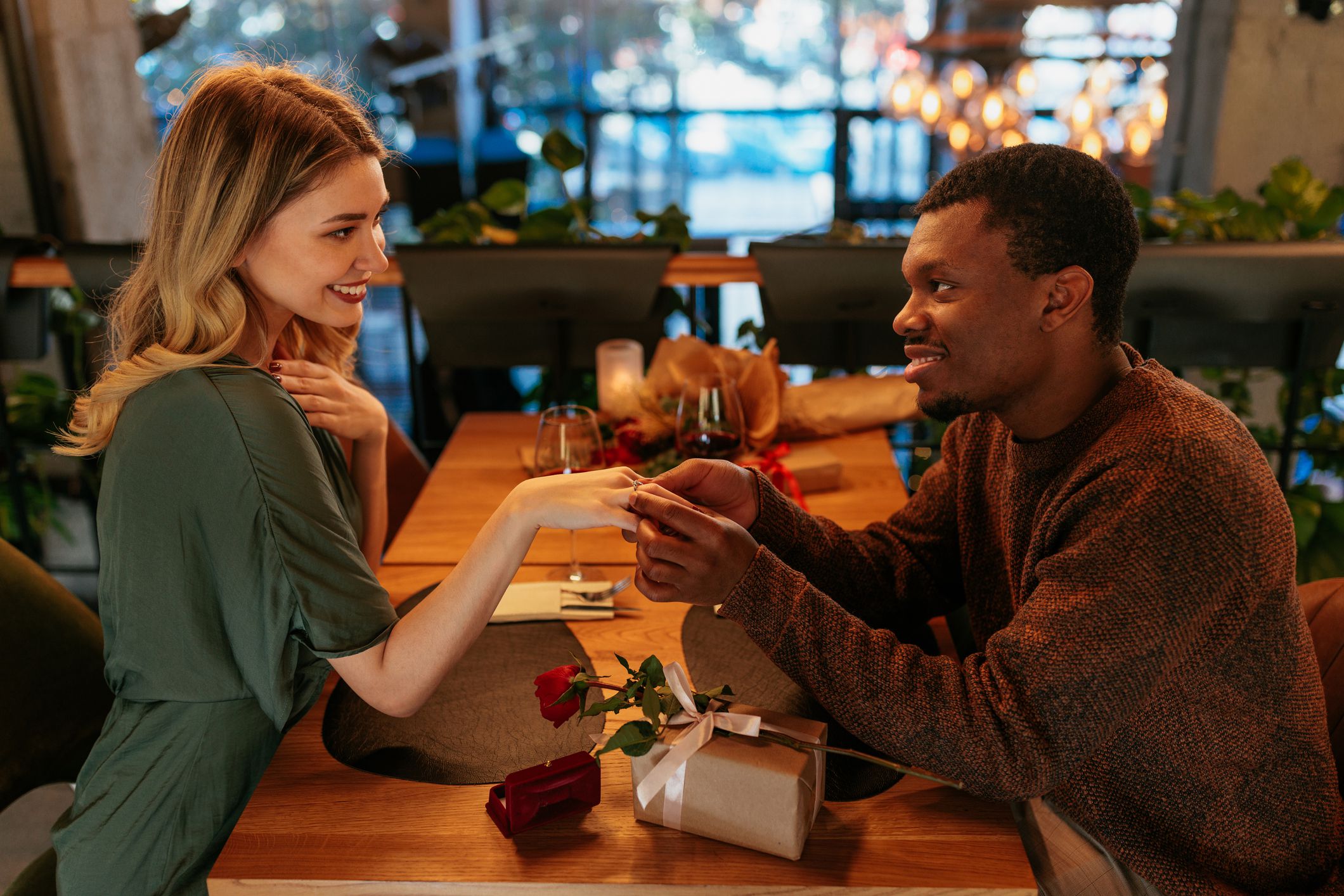 An African American man is putting an engagement ring on his Caucasian girlfriends finger on their date in a restaurant.
