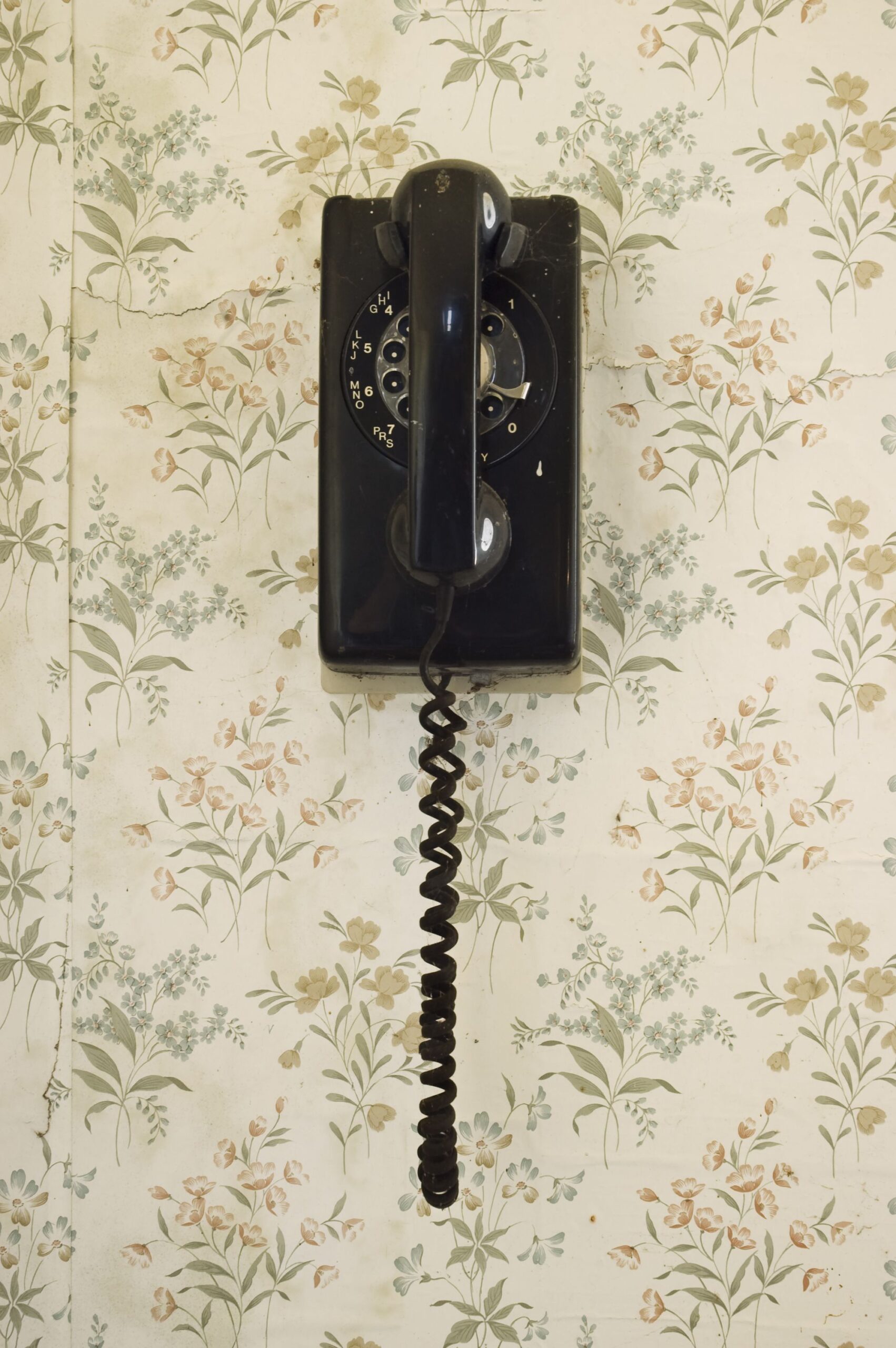 An old rotary dial phone hangs on the wall covered in faded flower wallpaper.