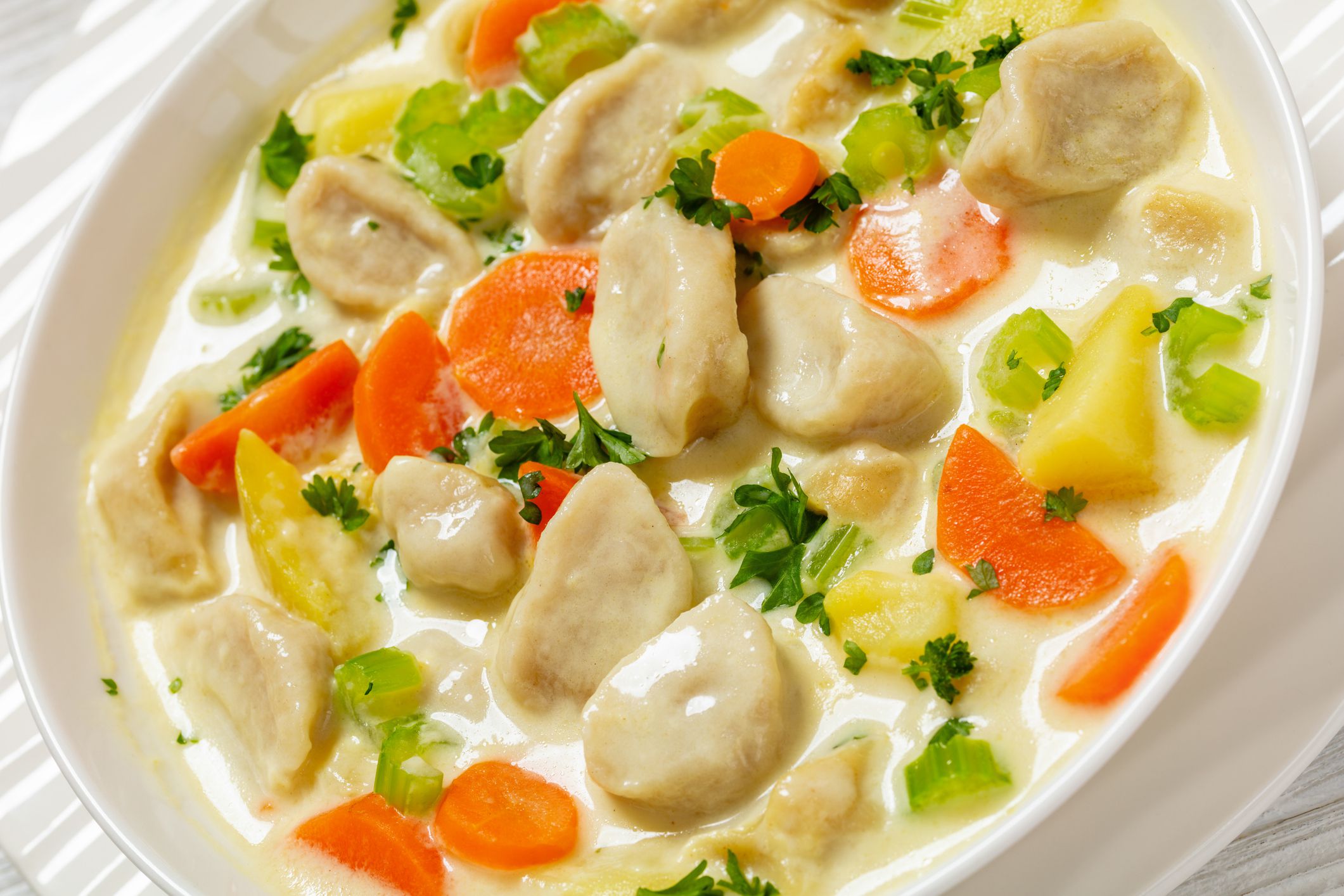 Knoephla soup, traditional german dumpling soup with potatoes, celery, carrots and parsley in white bowl on white wooden table, close-up, dutch angle view