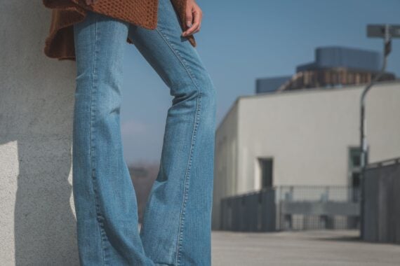A person wearing blue flared jeans and a brown sweater stands outdoors against a white wall, with buildings and a blue sky in the background.