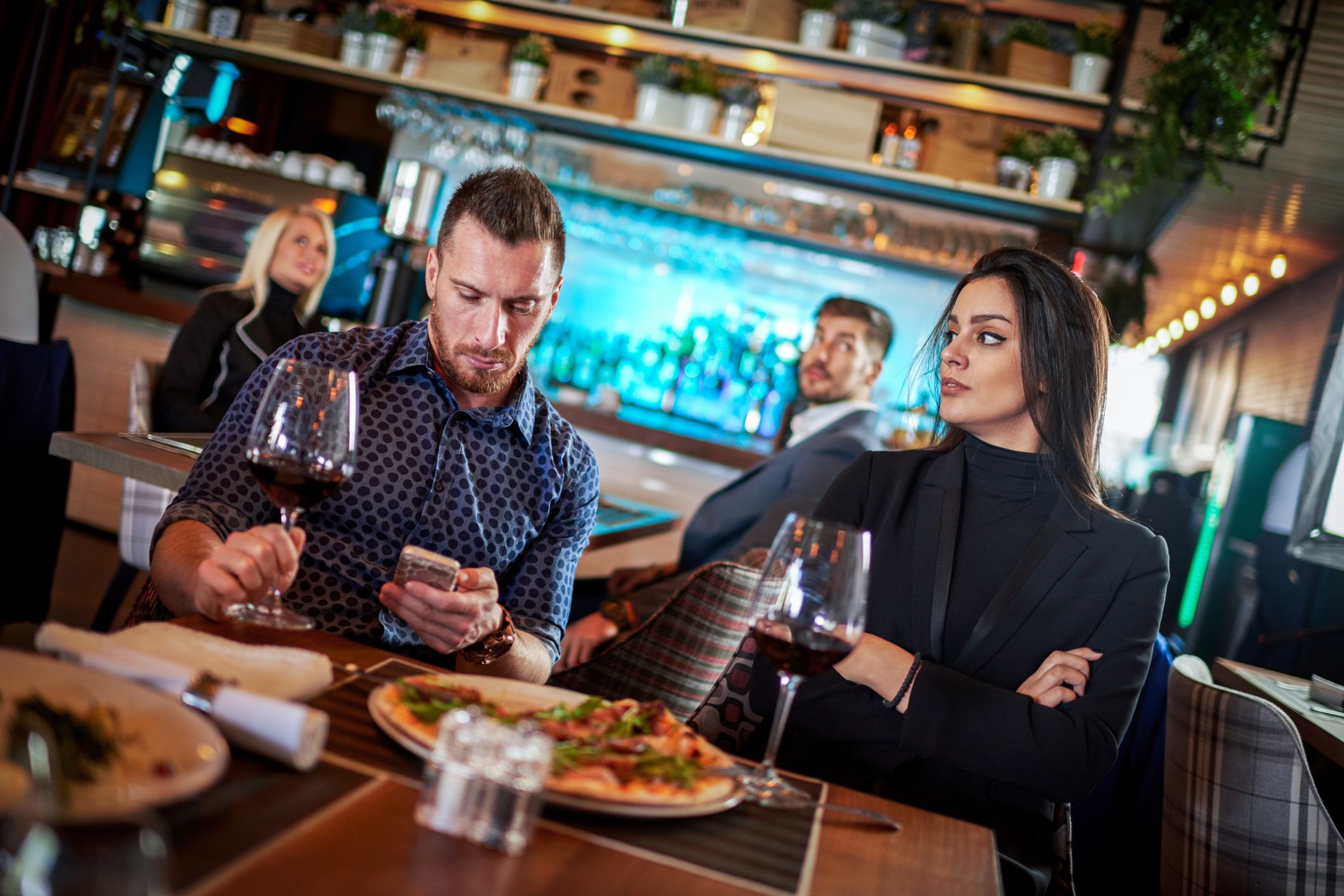 Modern heterosexual couple sitting in restaurant and woman is looking at her boyfriend while he is texting on mobile smart phone.