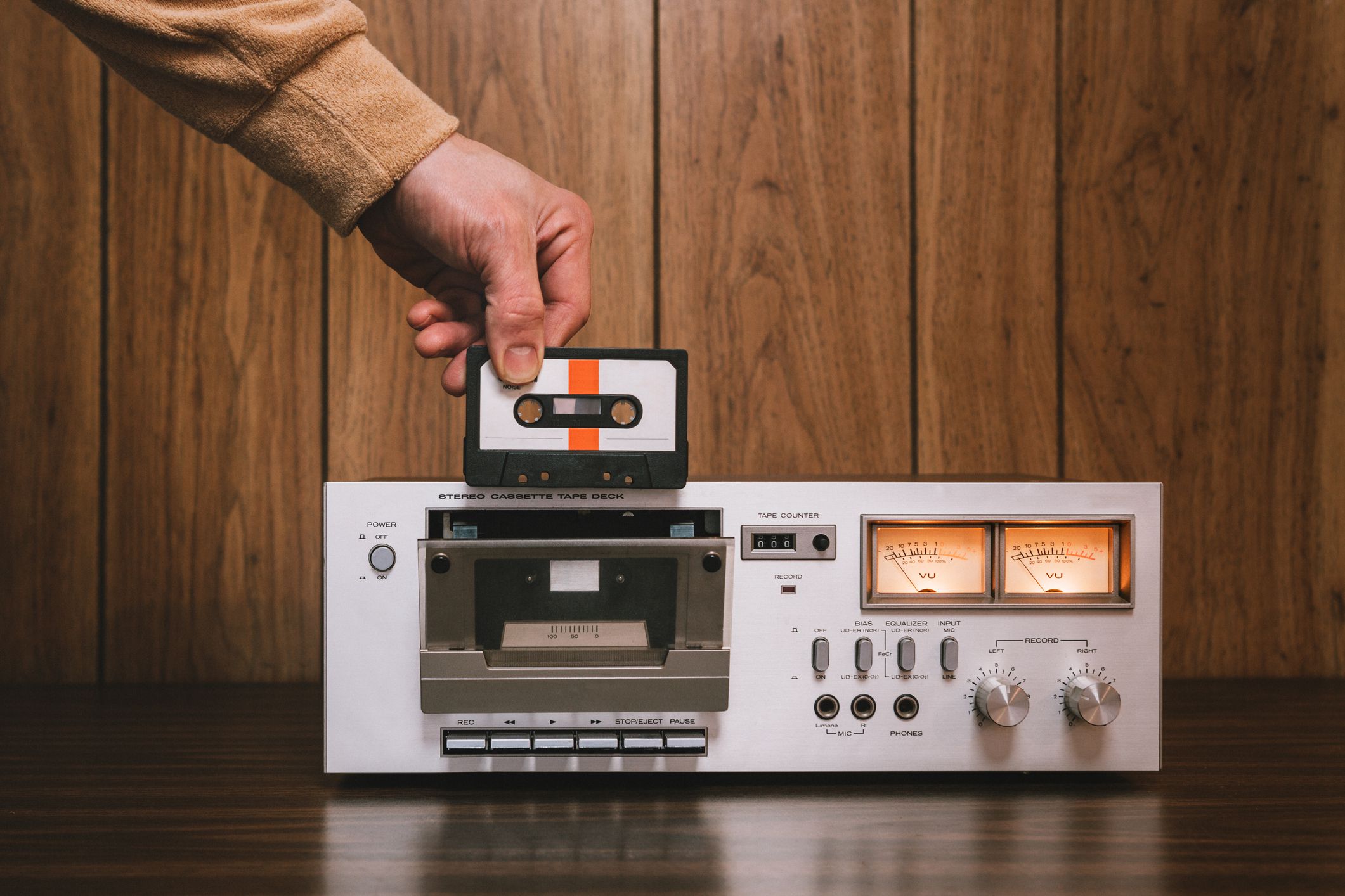 A vintage looking tape player / recorder stereo sits on the counter of a 1970's - 1980's living room with wood paneling. A mans hand reaches down to insert a cassette into the machine. Horizontal image with copy space.