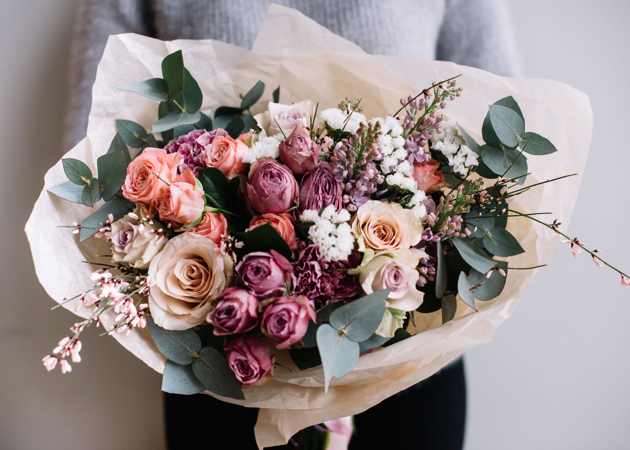 Young florist woman holding freshly made blossoming flower bouquet of roses and eucalyptus on the grey wall background.