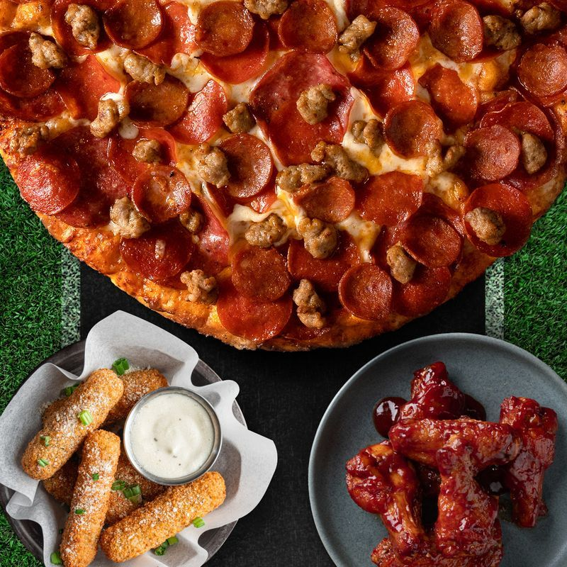 A pepperoni and sausage pizza, mozzarella sticks with dipping sauce, and saucy chicken wings are arranged on a dark surface next to green turf, resembling a football field.
