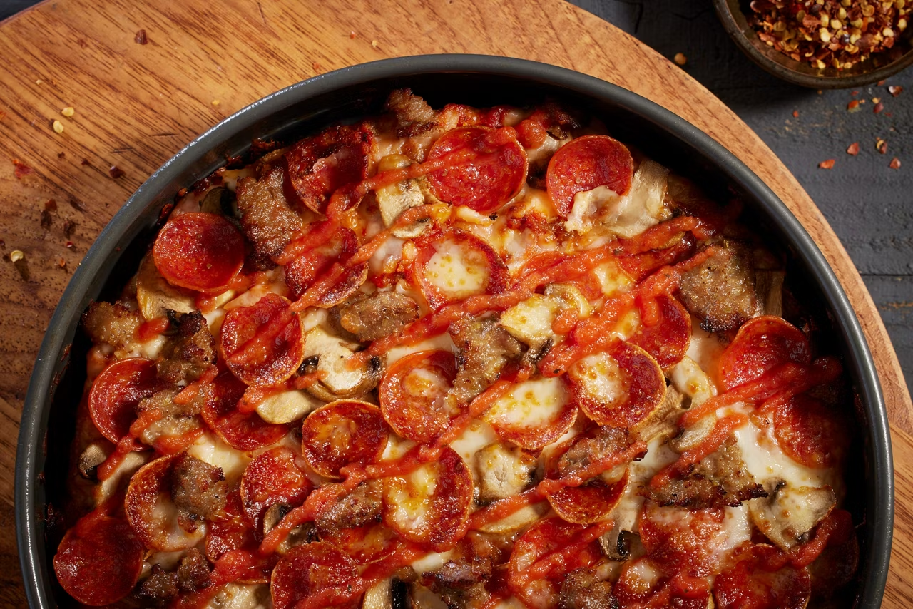 A close-up of a baked pizza topped with pepperoni, sausage, mushrooms, and melted cheese in a round black pan on a wooden board, with a bowl of red pepper flakes in the background.