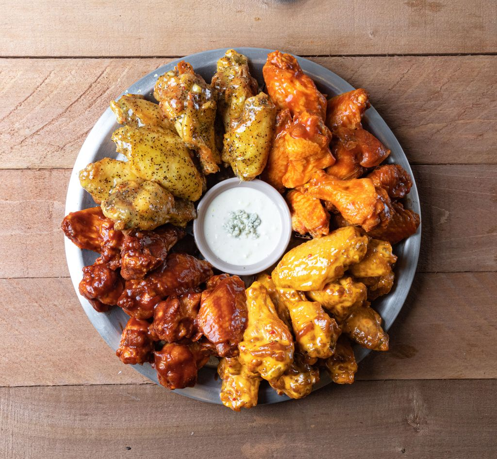 A round metal platter with four types of sauced chicken wings arranged in sections around a cup of ranch dipping sauce, placed on a wooden table.