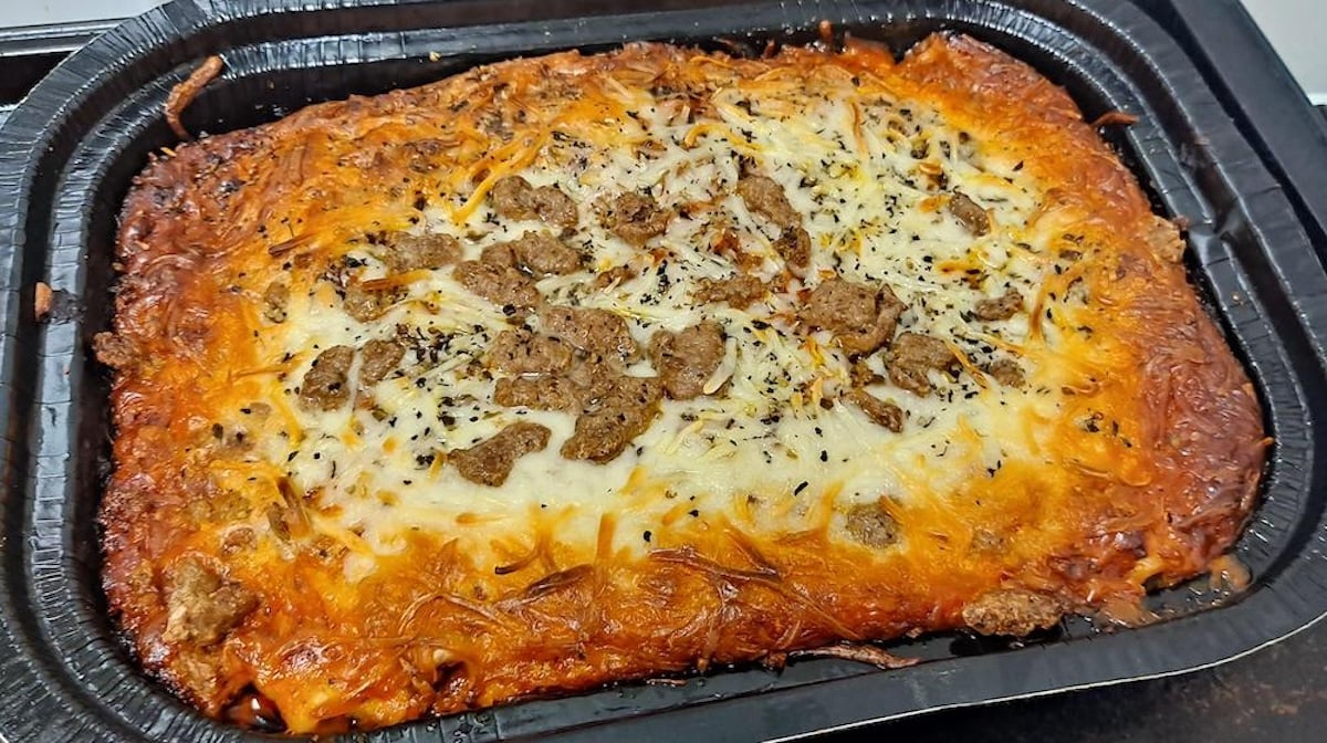 A rectangular baked dish in a black tray featuring a golden-brown, cheesy top with sprinkled herbs and pieces of browned meat, resembling a homemade lasagna or casserole.