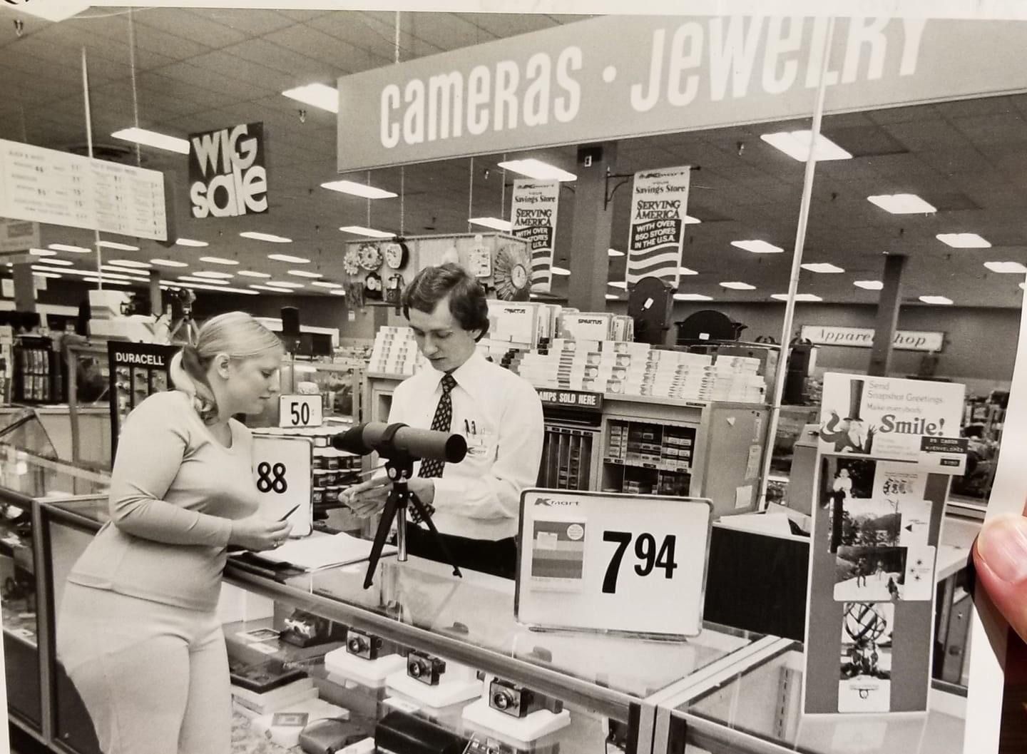 Man working at the camera department at a Kmart in the 70s helps a customer