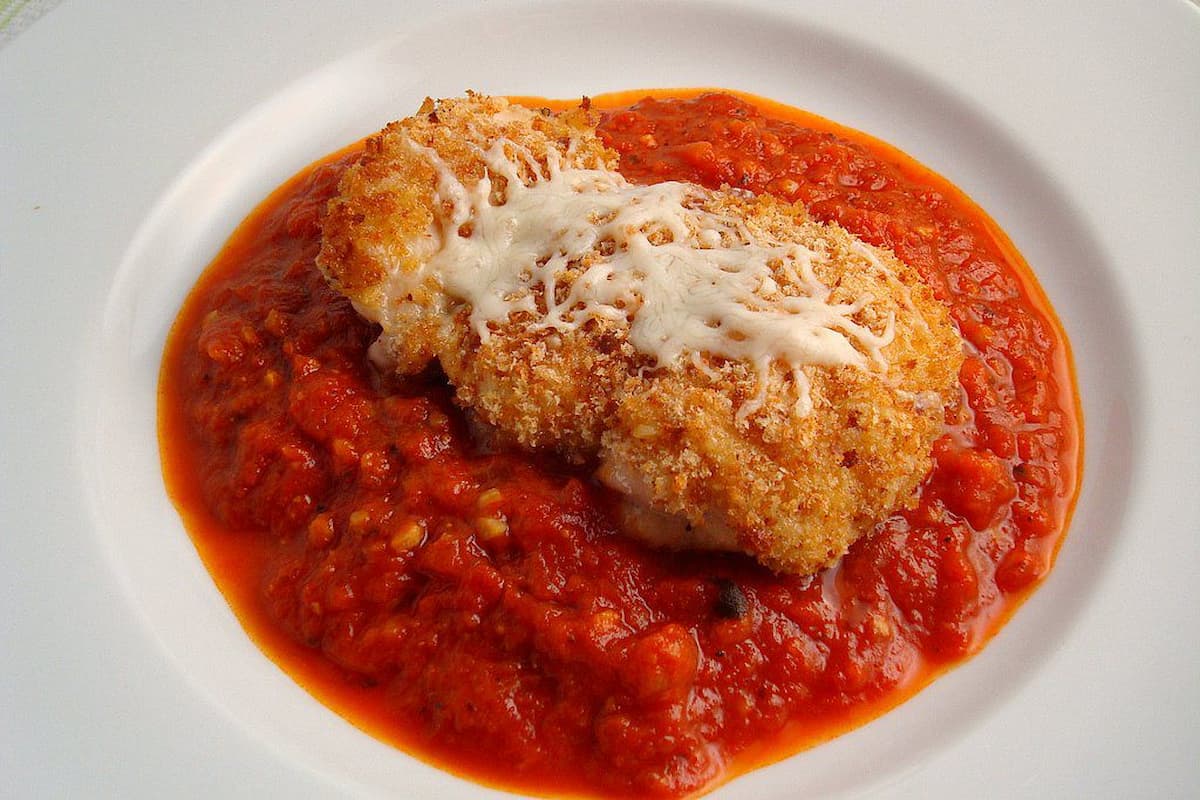 A breaded chicken breast topped with melted cheese sits on chunky red tomato sauce, served on a white plate—this popular takeout dish in every state is comfort food at its best.