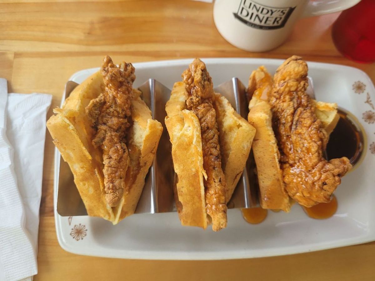 Three pieces of fried chicken are served inside folded waffles on a rectangular plate, with a cup in the background and syrup drizzled underneath one of the waffle sandwiches.