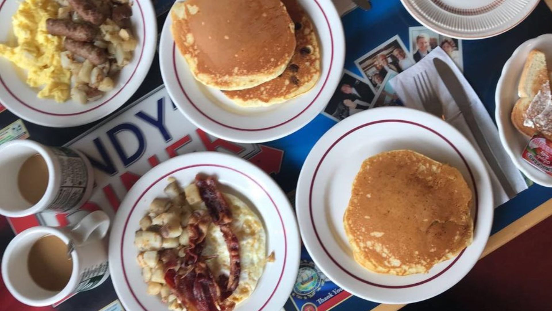 A table with plates of pancakes, scrambled eggs with sausage, bacon over hash browns, toast with jelly, and two mugs of coffee, set on a colorful placemat with photos and diner memorabilia.