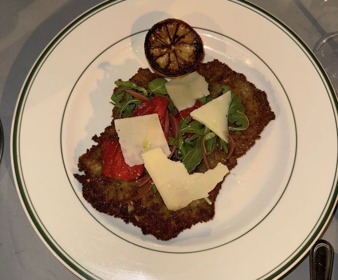 A breaded, pan-fried cutlet topped with arugula, sliced tomatoes, shaved cheese, and red onion, served with a grilled lemon half on a white plate with a green rim.