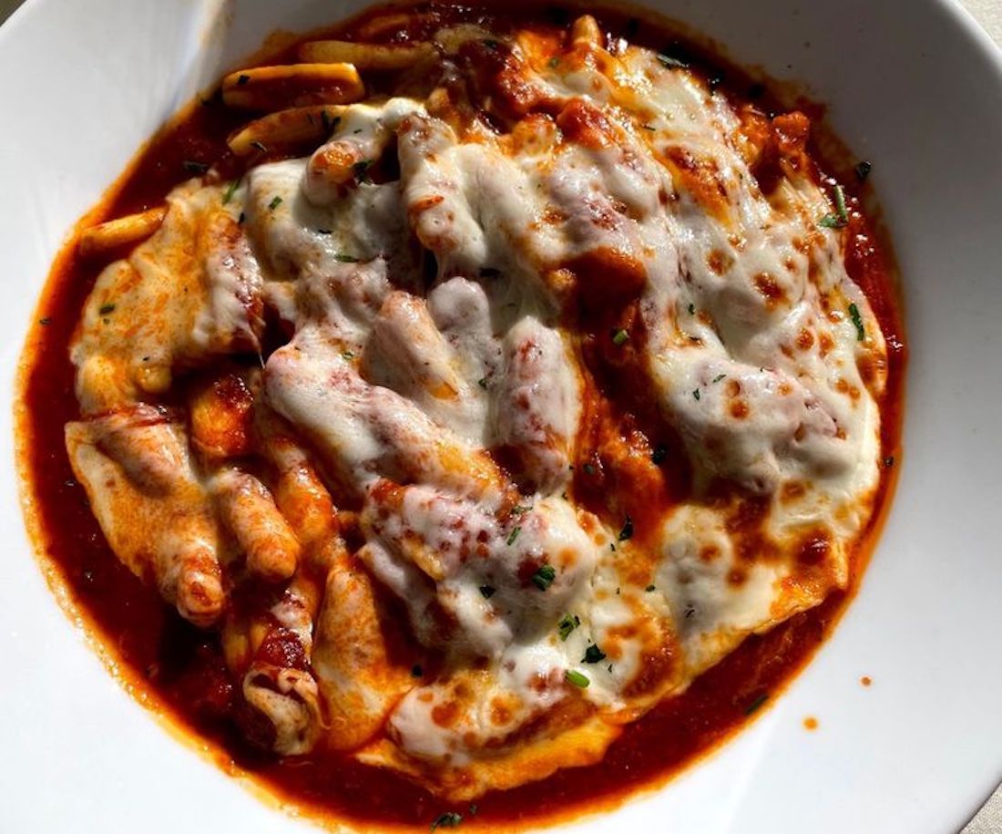Baked ziti pasta in tomato sauce, topped with melted mozzarella cheese and sprinkled with herbs, served in a white bowl.