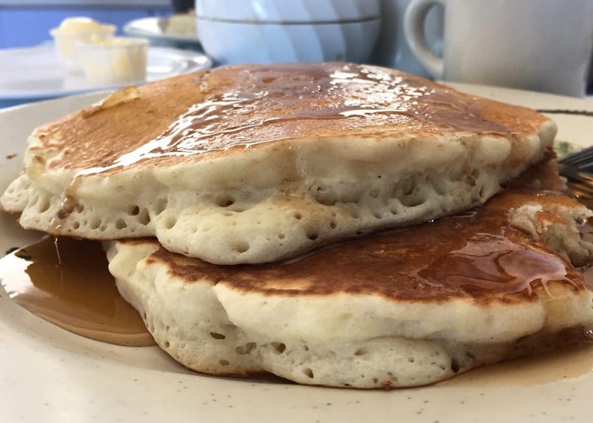 Two fluffy pancakes stacked on a plate, topped with syrup. A fork is beside the pancakes, and a coffee cup and butter are visible in the blurred background.