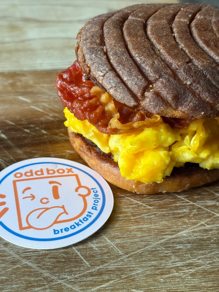 A breakfast sandwich with scrambled eggs and crispy bacon on a round toasted bun sits on a wooden surface next to a circular sticker labeled "oddbox breakfast project" with a quirky face illustration.