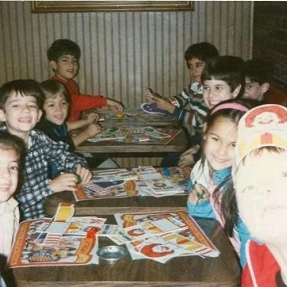 Eight children are sitting in two booths at a restaurant, smiling at the camera. They have paper placemats and colorful items on the tables. One child in the foreground is wearing a hat and is partially overexposed by the camera flash.