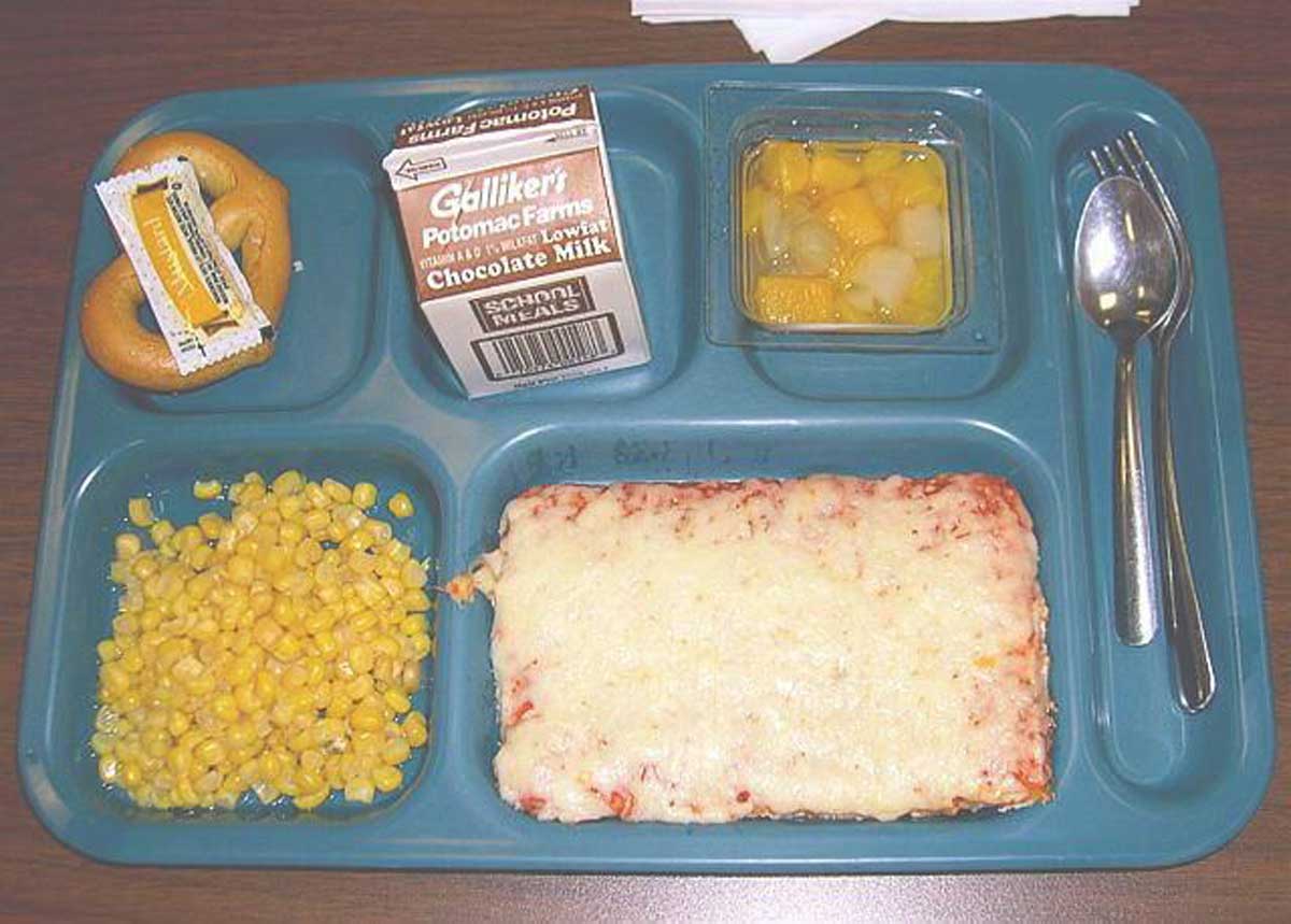 A blue cafeteria tray displays a classic school lunch—cheese pizza, corn, mixed fruit, a soft pretzel with mustard, and chocolate milk—reflecting popular choices as school lunch prices by year continue to change.
