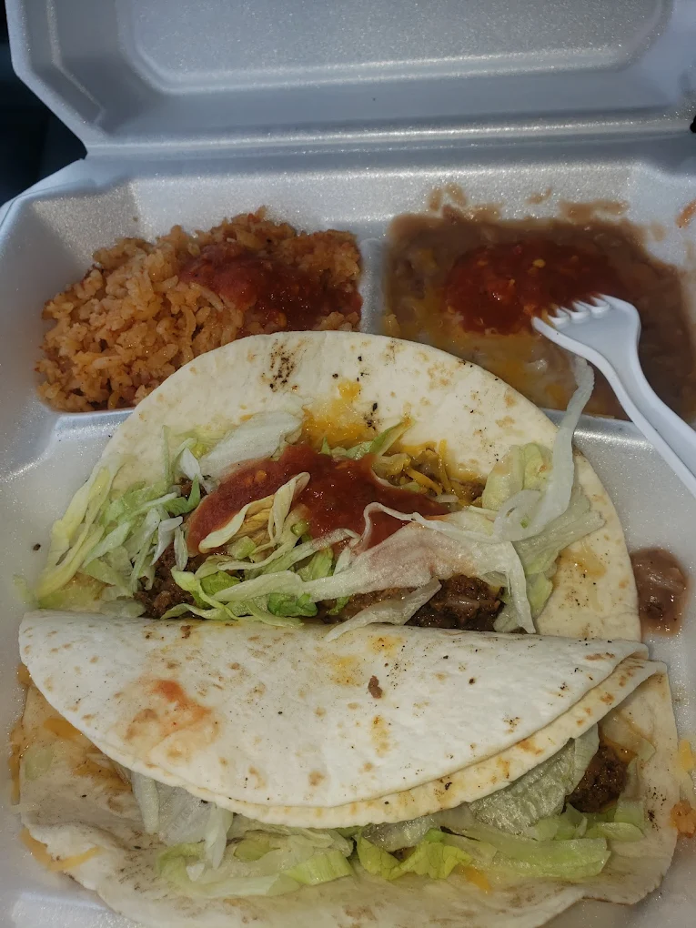 A takeout container with two soft tacos filled with ground beef, lettuce, cheese, and salsa, served with a side of Mexican rice and refried beans topped with salsa. A white plastic fork is in the beans.