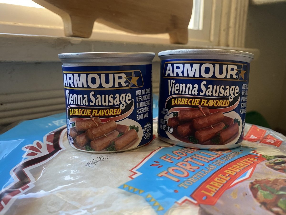 Two cans of Armour Vienna Sausage, barbecue flavored, sit side by side on a package of flour tortillas near a window. The cans feature images of sausages and blue and yellow branding.