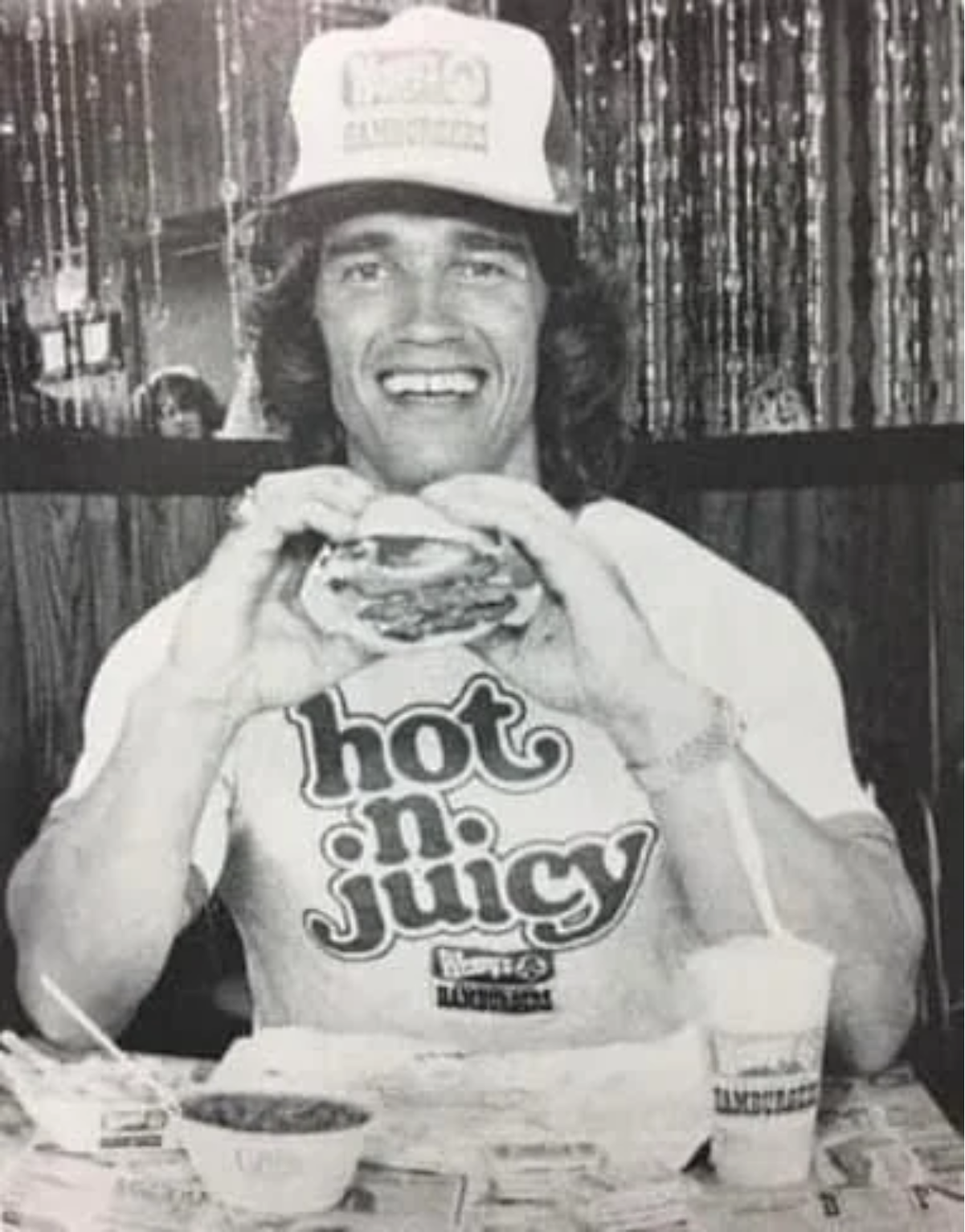 A man wearing a "hot n. juicy" t-shirt and a branded cap smiles while holding a hamburger. He sits at a table with a cup and a bowl of food in front of him, with beaded curtains in the background.