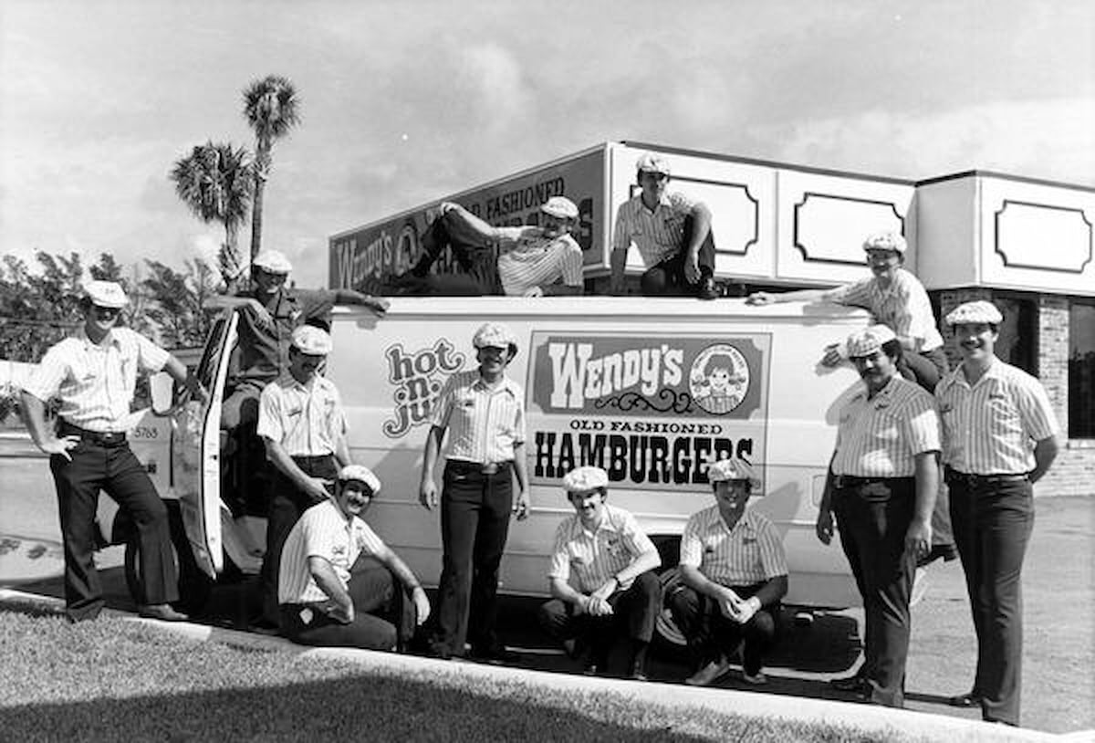 A group of Wendy’s employees in uniform pose around a delivery van labeled “Wendy’s Old Fashioned Hamburgers” outside a restaurant, capturing the charm of vintage Wendy’s photos with palm trees and a partly cloudy sky in the background.