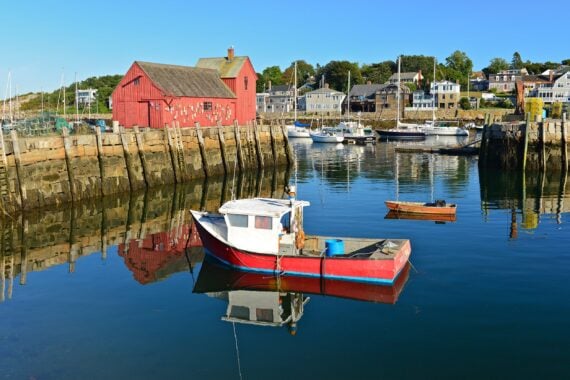 A small red and white fishing boat floats in calm harbor waters near a large red building, with other boats and waterfront houses in the background under a clear blue sky.