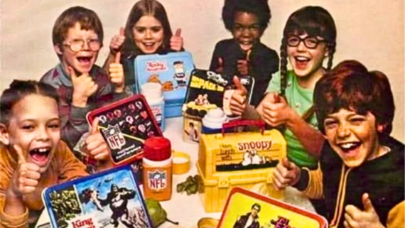 Six children sit at a table, smiling and giving thumbs up, with colorful vintage lunchboxes and thermoses featuring cartoon and movie characters in front of them.
