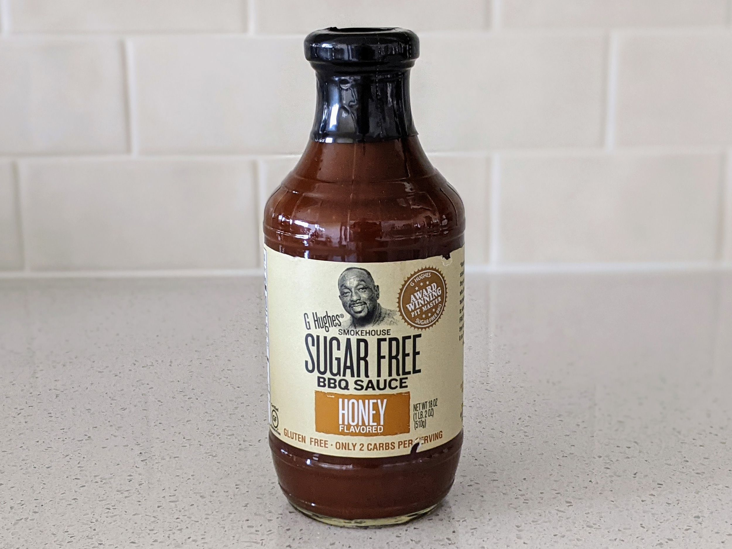 A Bottle of G. Hughes Smokehouse Sugar Free Honey BBQ Sauce on a beige countertop, beige kitchen wall tile in the background