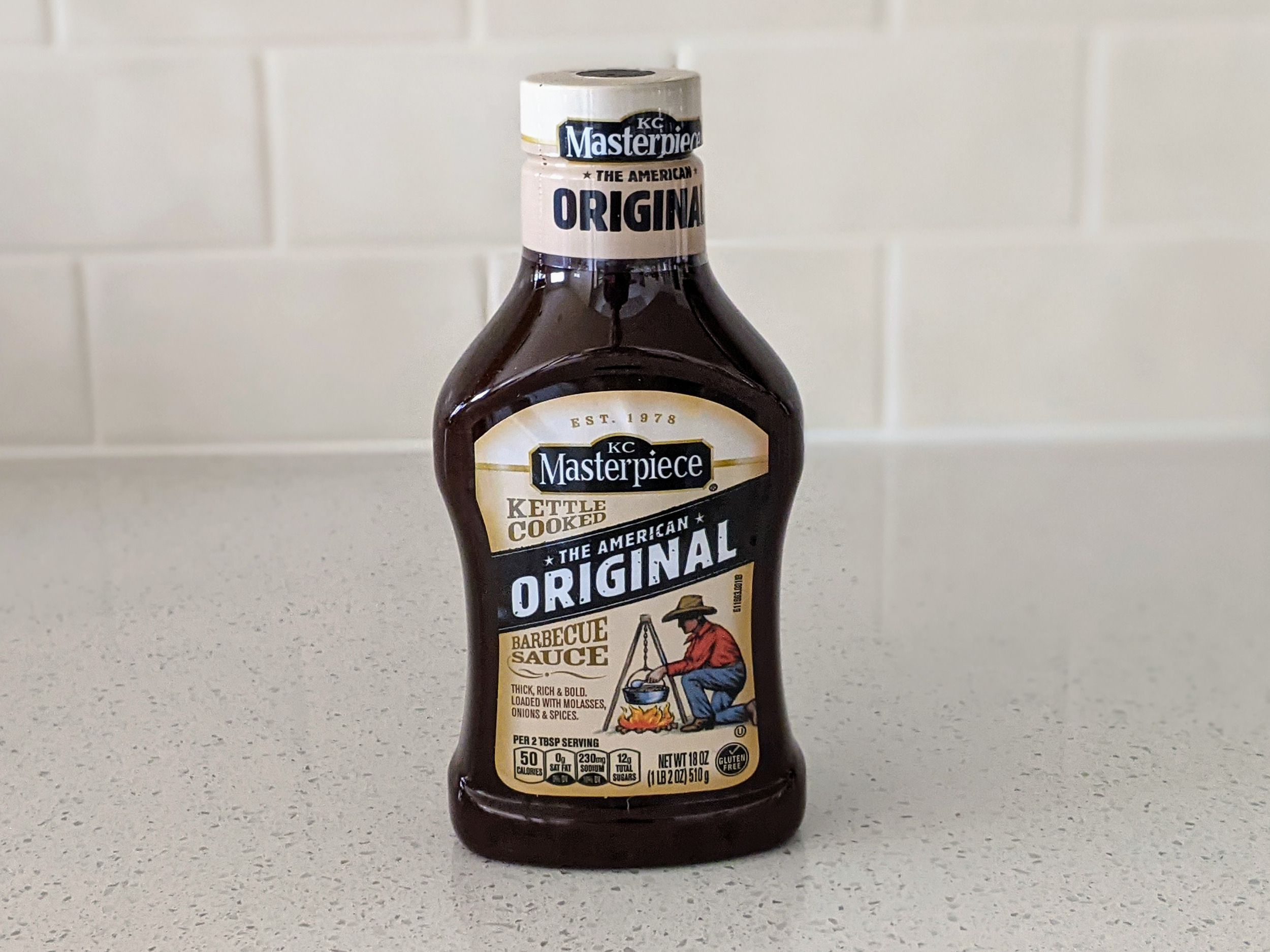 A Bottle of KC Masterpiece American Original Barbecue Sauce on a beige countertop, beige kitchen wall tile in the background