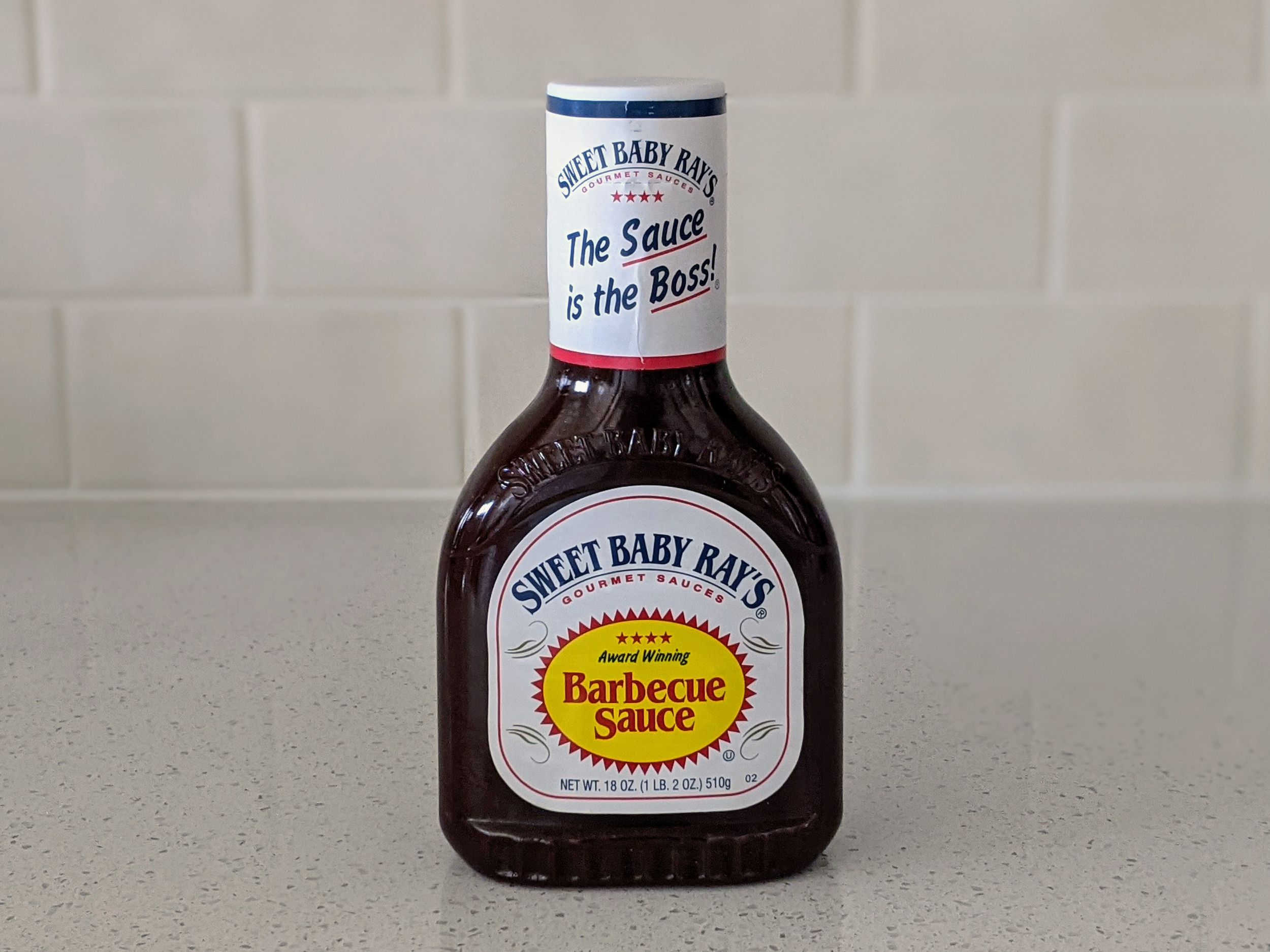 A Bottle of Sweet Baby Ray’s Barbecue Sauce on a beige countertop, beige kitchen wall tile in the background