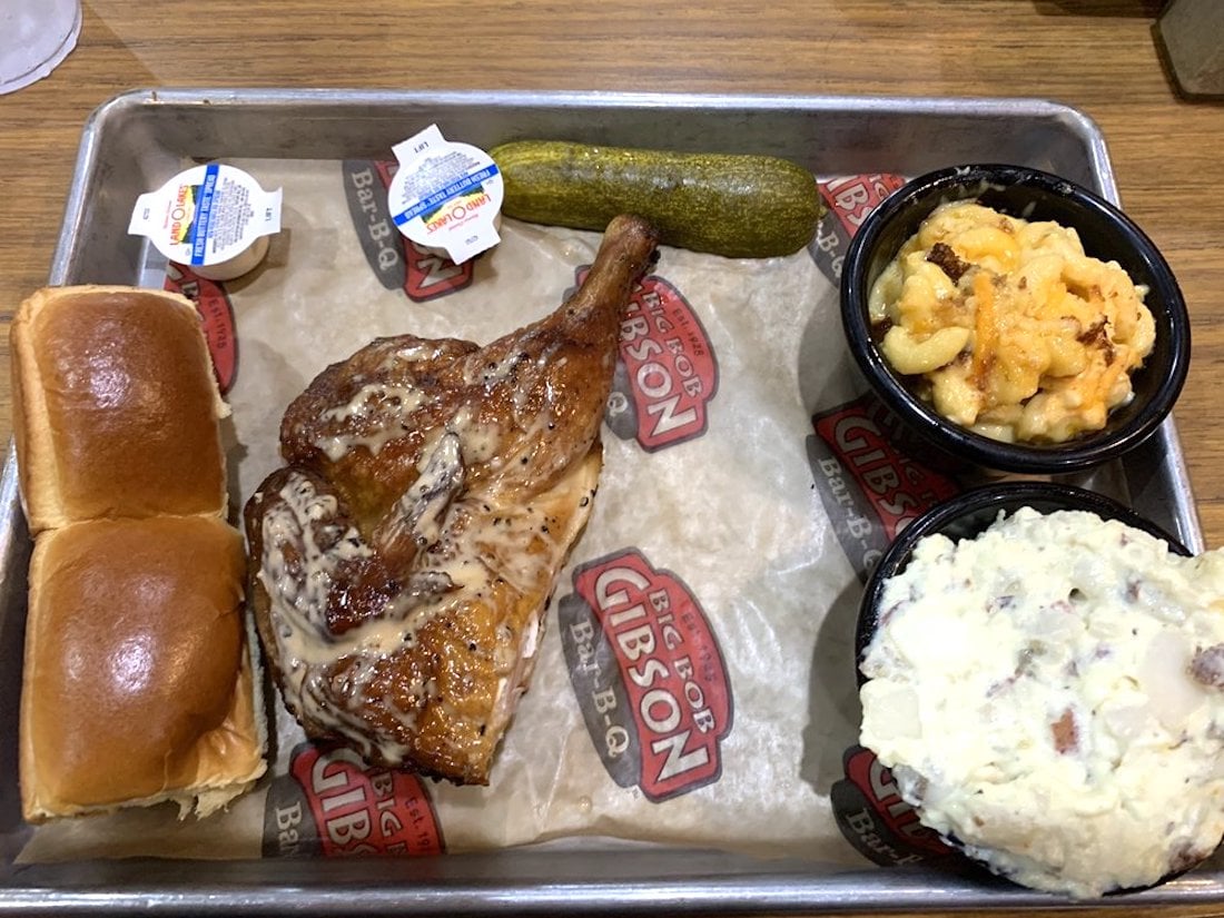 A tray with a grilled chicken leg quarter, two dinner rolls with butter, a pickle, mac and cheese, and potato salad, all served on butcher paper with "Gibson BBQ" logos.