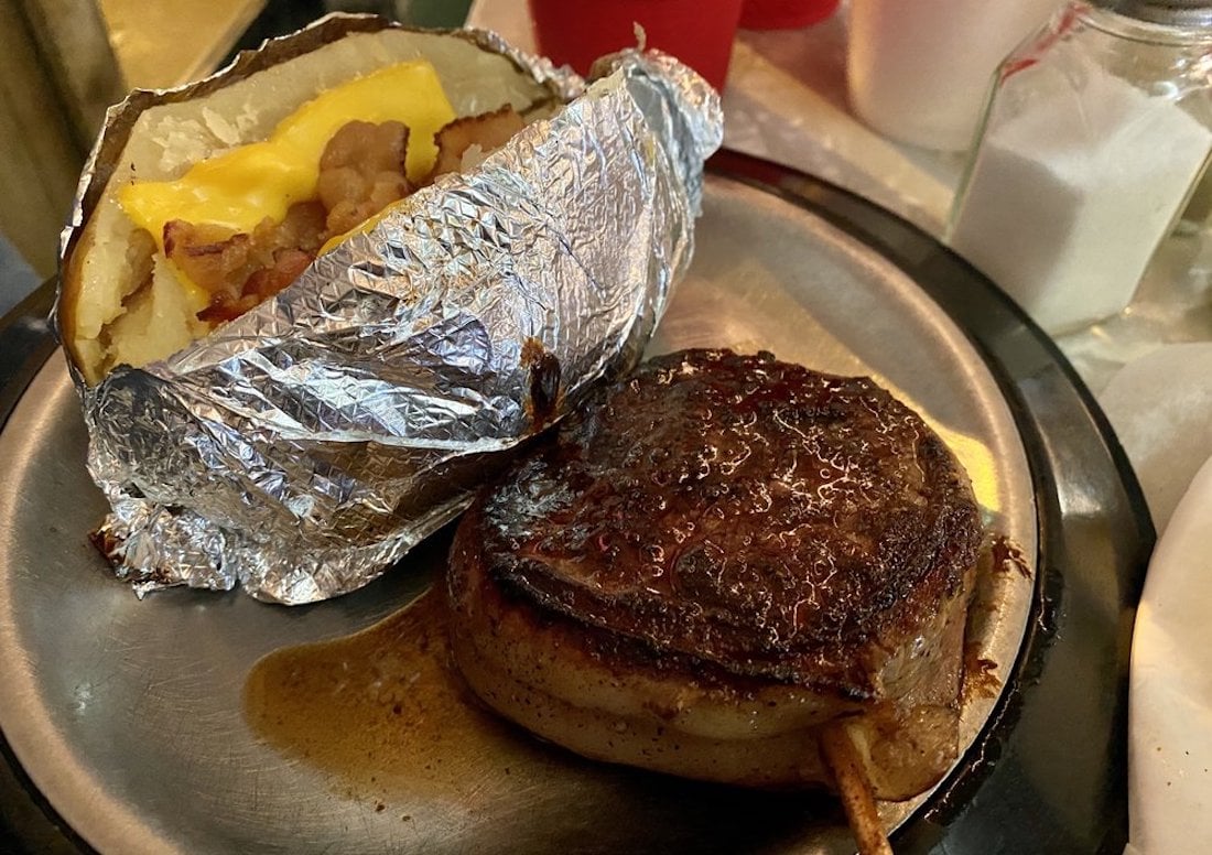 A cooked filet mignon wrapped in bacon sits on a metal plate next to a baked potato topped with melted cheese and bacon bits, partially wrapped in foil. A salt shaker is in the background.