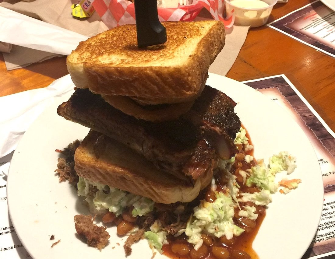A large sandwich with stacked toasted bread, ribs, onion rings, and barbecue meat on a white plate with coleslaw and baked beans, with a knife stuck through the middle.