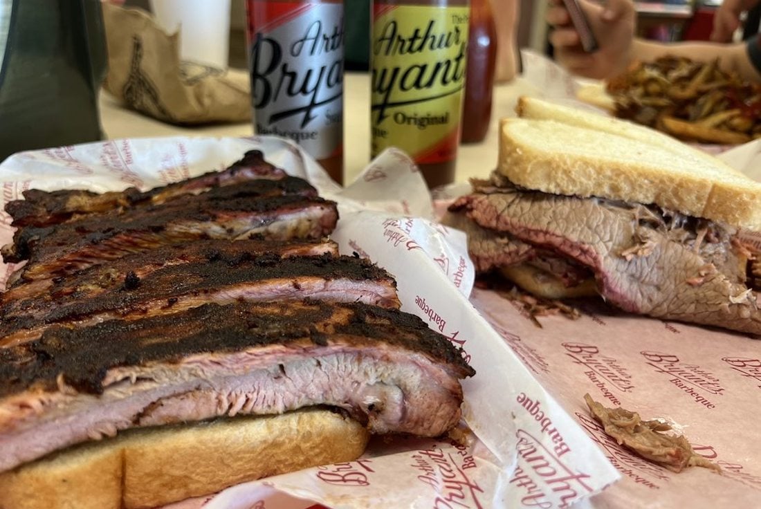 A close-up of barbecue food, featuring a rack of grilled ribs, a beef brisket sandwich on white bread, and two cans of Arthur Bryant’s barbecue sauce in the background.